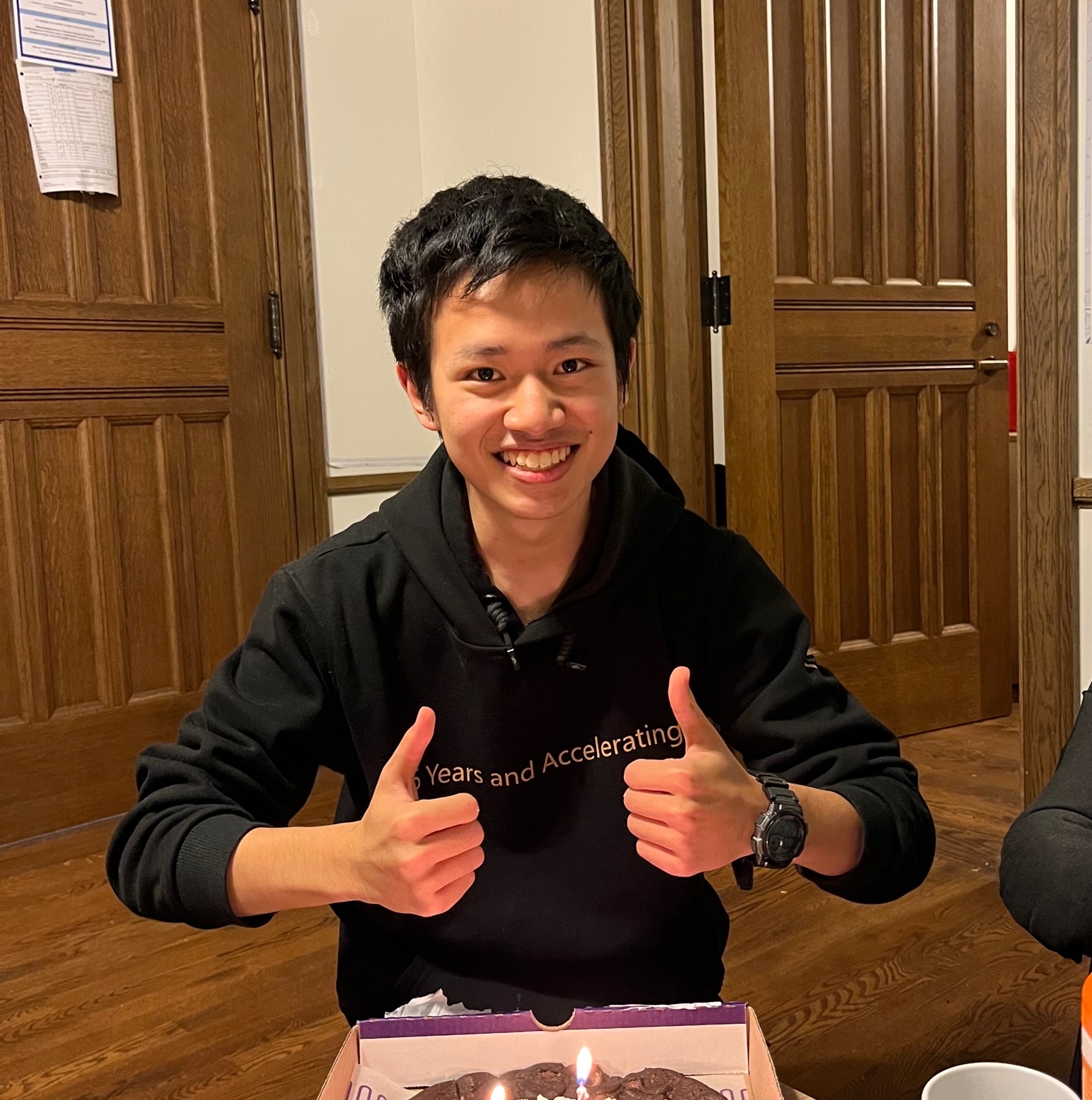 A man seated at a table, smiling and giving a thumbs up beside a cake with lit candles on them.